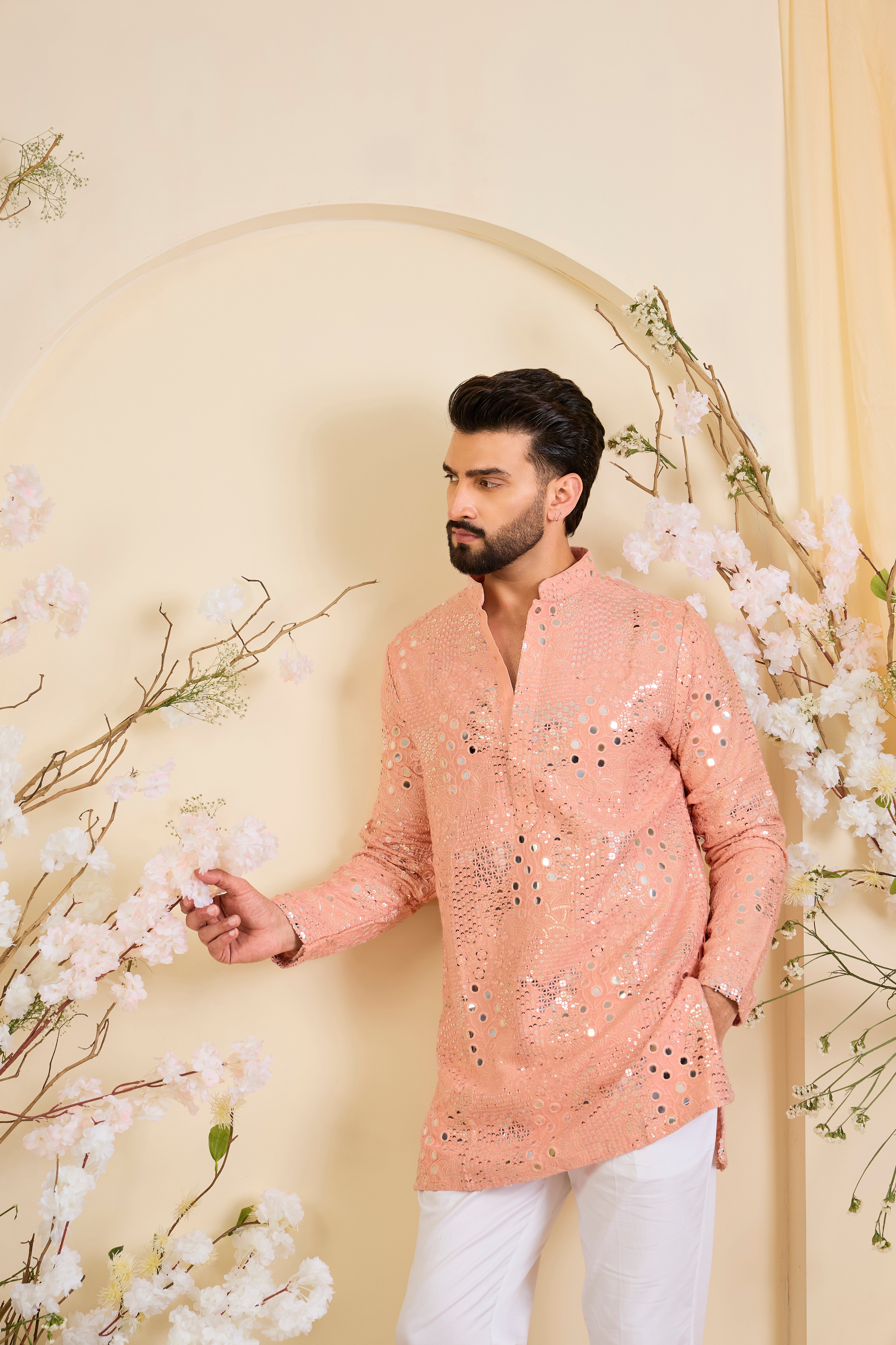 A peach short kurta enriched with intricate mirror work, delicate floral embroidery, and dense sequin detailing.